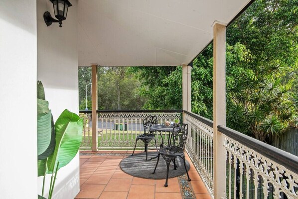 A covered balcony with a table and chairs, surrounded by a decorative railing and lush greenery.