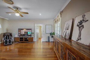 A spacious living room with a wooden floor, a ceiling fan, a massage chair, a flat-screen TV, and artwork on the walls.