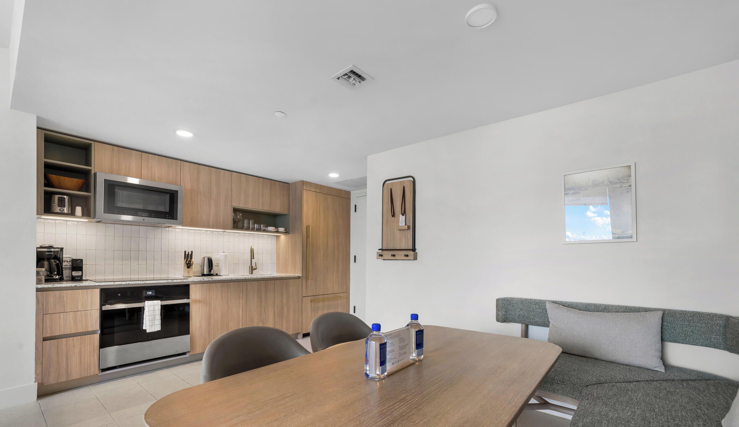 A modern kitchen with wooden cabinets, a microwave, and a built-in oven. There is a dining table with chairs and a sofa with a cushion.