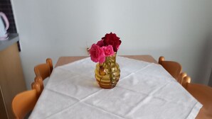 Ein Tisch mit weißer Tischdecke, eine gelbe Vase mit rosa Blumen und Holzsessel.