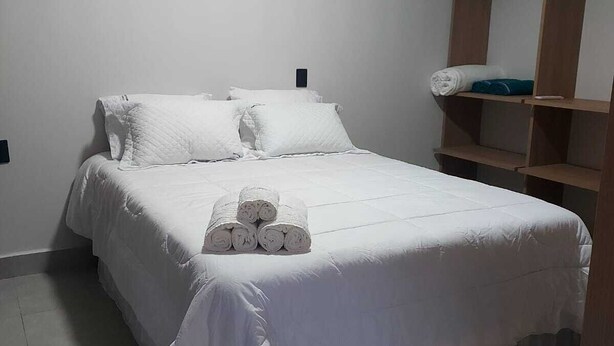 Une chambre avec un lit simple, recouverte d’une courtepointe blanche, avec des oreillers et des serviettes enroulées posés sur le lit. Une étagère en bois se trouve à droite du lit.