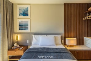 Quarto de hotel com cama, mesa de cabeceira, luminária, guarda-roupa e duas fotos emolduradas na parede.