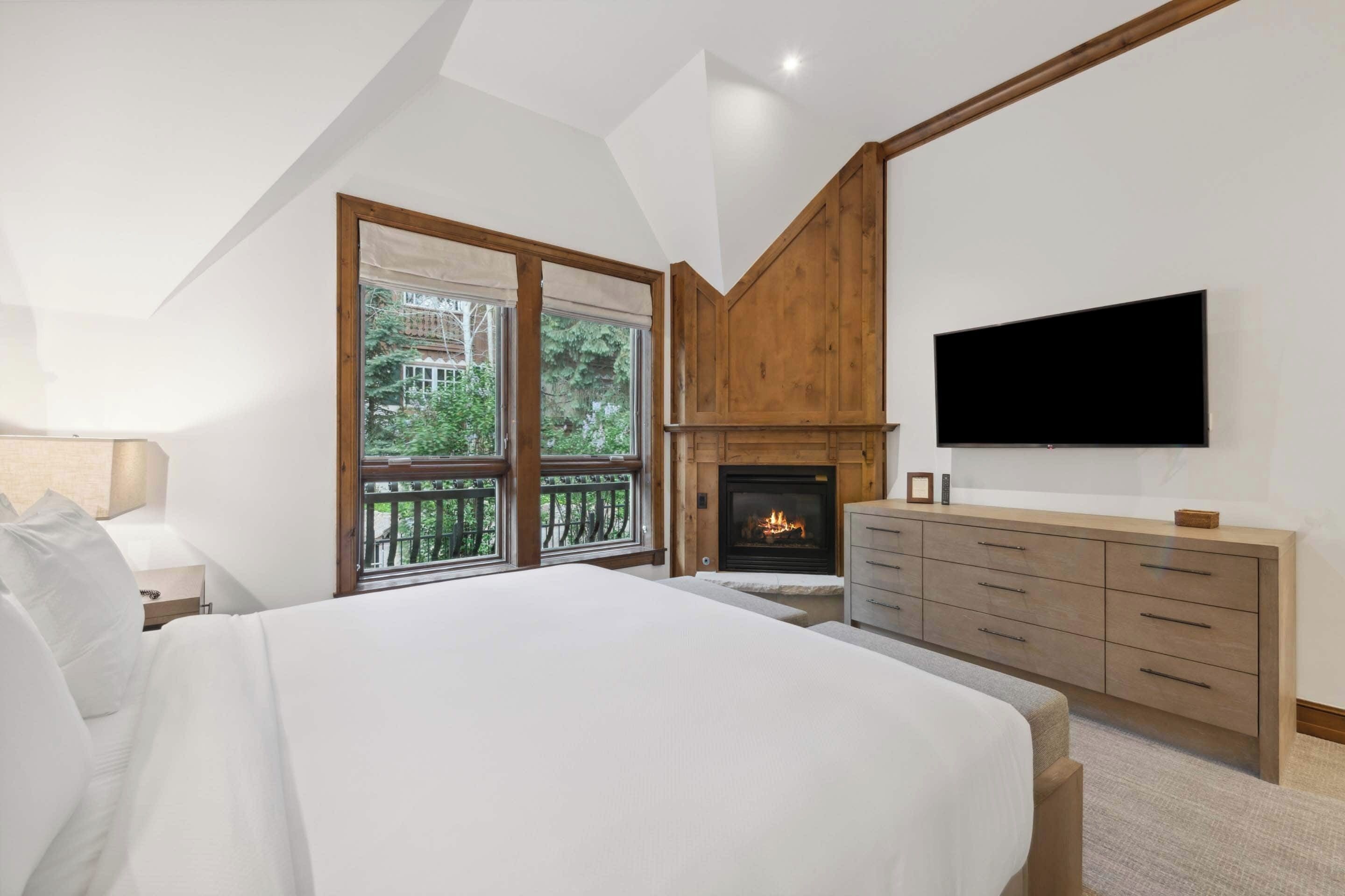 Een slaapkamer met een groot bed, een open haard, een aan de muur bevestigde televisie en een balkon met uitzicht op groen.