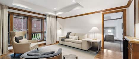 A spacious living room with a sofa, armchair, and a coffee table. There is a wooden cabinet and a lamp on a side table.