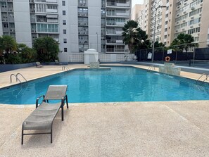 Ein Swimmingpool mit zwei Liegestühlen, umgeben von einem Zaun, mit Hochhäusern im Hintergrund.