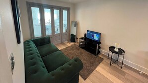 Uma sala de estar com um sofá verde, uma televisão de tela plana, uma mesinha com um abajur e uma janela grande com vidro fosco.
