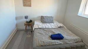 Quarto com cama, mesa de cabeceira com abajur, janela e uma frase motivacional na parede.