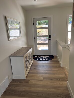 Un couloir avec un plancher en bois, une porte blanche dotée d’une fenêtre, un paillasson et un tableau encadré accroché au mur.