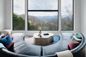 A modern living room with a curved black leather sofa, a round coffee table, and a view of a mountainous landscape through large windows.