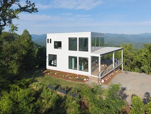 A modern two-story house with large windows, a deck, and a view of the surrounding forest and mountains. - The Heartscape Collection | 3 Private Mountain Homes Near Asheville (Marshall)