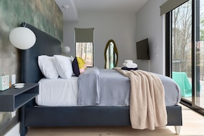 A modern bedroom with a large bed, a bedside table, a wall-mounted TV, and a window with a view of trees. - The Heartscape Collection | 3 Private Mountain Homes Near Asheville (Marshall)