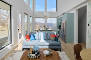 A modern living room with a sofa, a coffee table with books, and a view of mountains through large windows. - The Heartscape Collection | 3 Private Mountain Homes Near Asheville (Marshall)