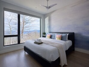 A bedroom with a large window, a bed with pillows, a bedside table, and a ceiling fan.