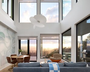 A modern interior with a dining area, a sofa, and large windows offering a view of the outdoors. - The Heartscape Collection | 3 Private Mountain Homes Near Asheville (Marshall)