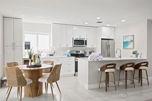 Una cocina moderna con muebles blancos, electrodomésticos de acero inoxidable, una isla central con taburetes y un comedor con sillas de mimbre.