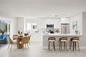 Una cocina moderna con armarios blancos, electrodomésticos de acero inoxidable, una isla central con taburetes y un comedor con sillas de mimbre.
