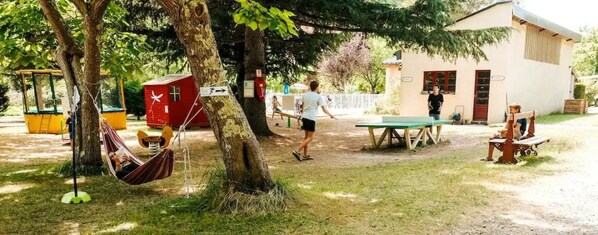 A grassy outdoor area with a swing set, a hammock, a red playhouse, and a table tennis table.