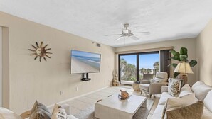 A living room with a ceiling fan, a flat-screen TV, a large window with a view of palm trees, and a seating area with a coffee table and sofas. - Las Brisas 521 (New Smyrna Beach)