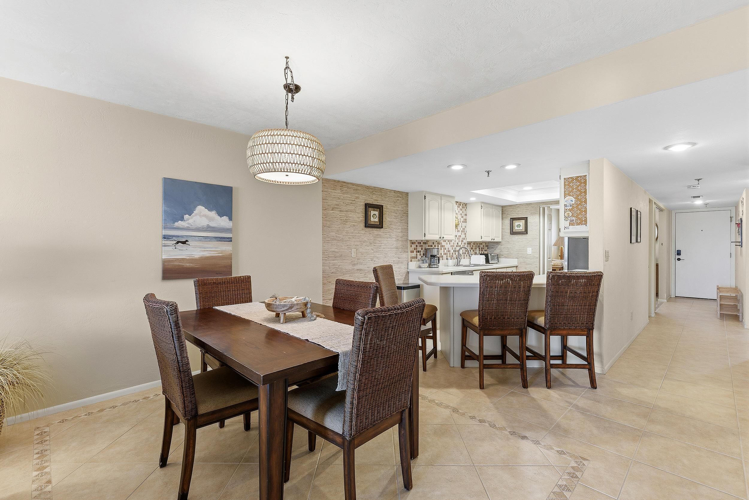 A dining area with a wooden table and wicker chairs, a kitchen with white cabinets, and a hallway leading to a door.