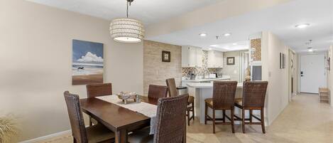 A dining area with a wooden table and wicker chairs, a kitchen with white cabinets, and a hallway leading to a door.