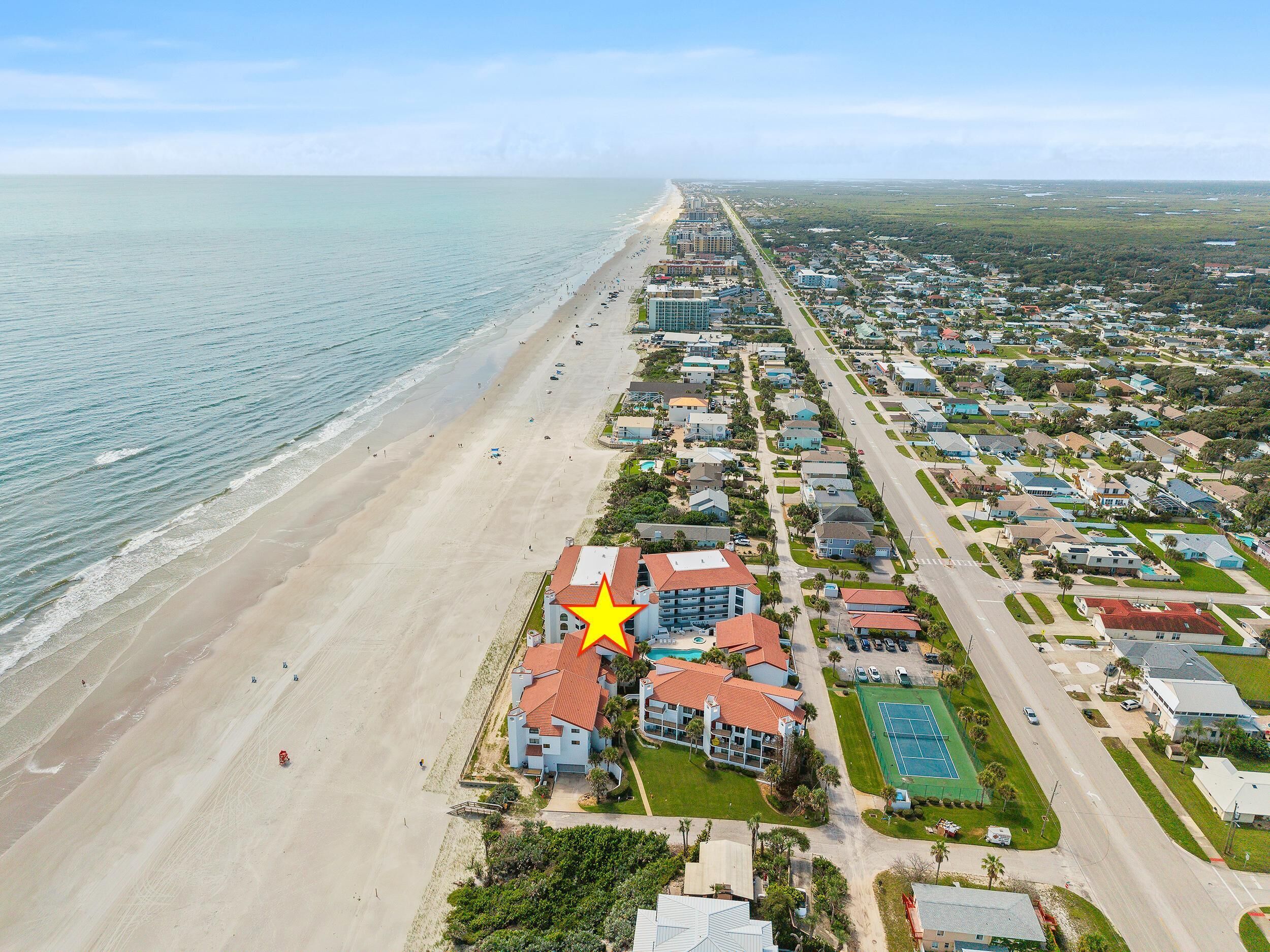Vista aérea de una zona residencial costera con propiedades frente a la playa, una carretera paralela a la playa y un edificio señalado con techo rojo.