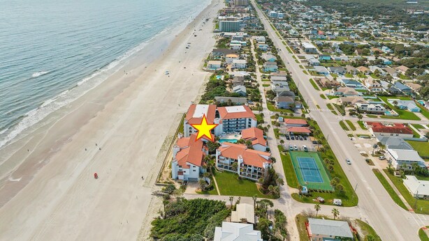 An aerial view of a coastal residential area with beachfront properties, a road parallel to the beach, and a marked building with a red roof. - Las Brisas 521 (New Smyrna Beach)