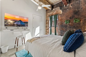 A bedroom with a bed, a desk, a chair, a television, and a brick wall. - 2BR Modern Retreat Escape | Downtown Memphis | Walking Distances | Arcade & Gym (Memphis)