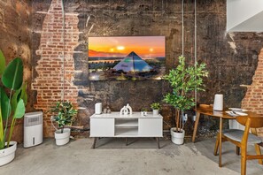 Un soggiorno moderno con parete in mattoni, una TV a parete che mostra una vista al tramonto, un tavolino consolle bianco e piante in vaso.