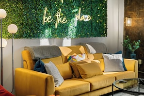 A yellow sofa with multiple cushions, a green wall with a 'be the place' sign, and a floor lamp. - 2BR Modern Retreat Escape | Downtown Memphis | Walking Distances | Arcade & Gym (Memphis)