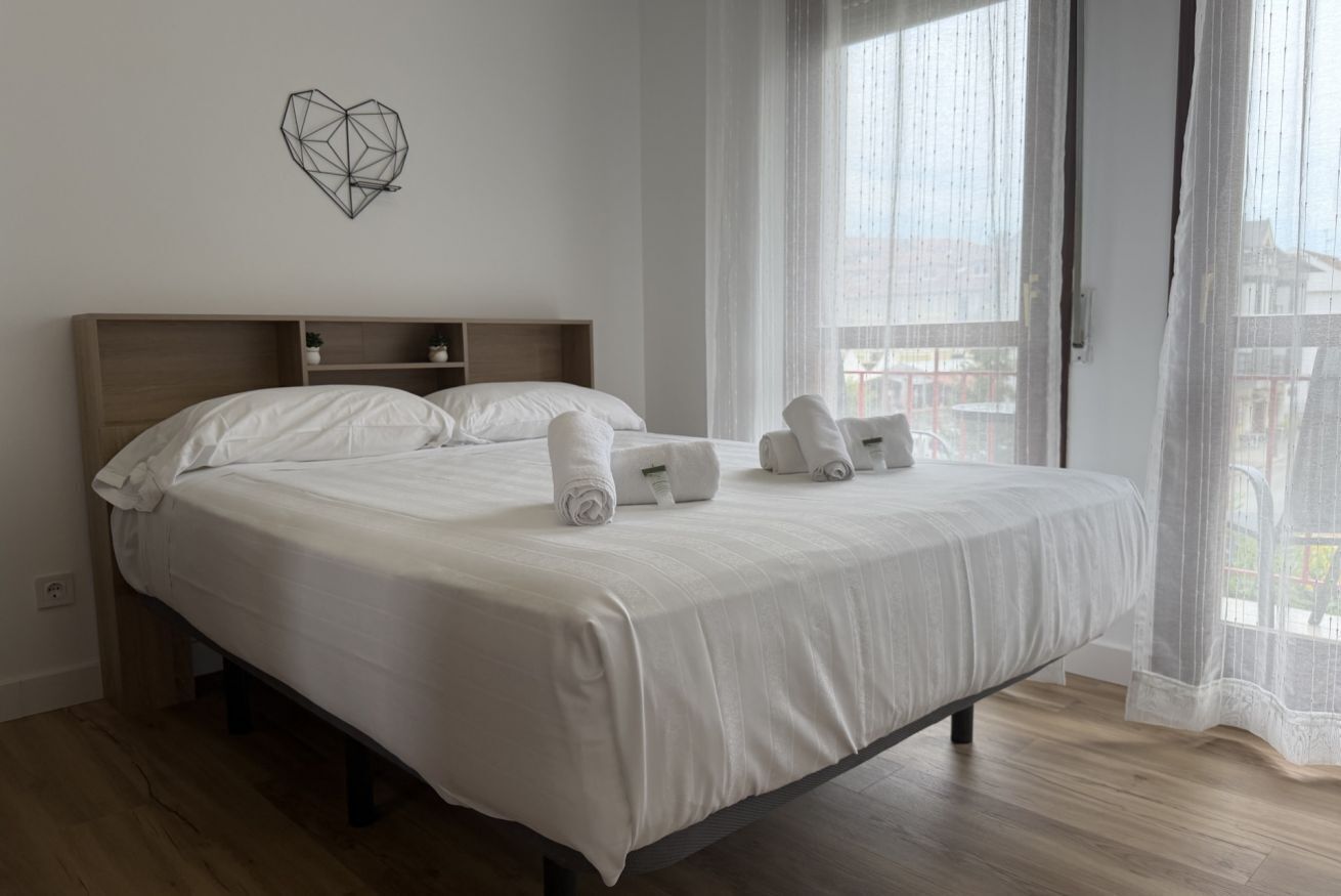 A neatly made bed with white linens and towels, a geometric wall decoration, and a view through sheer curtains.