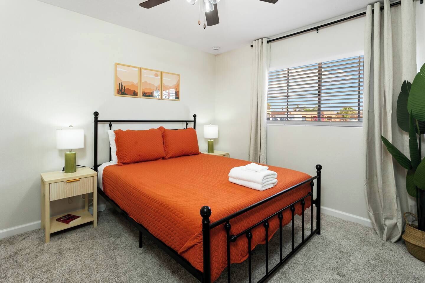 A neatly arranged bedroom with a bed, bedside tables, a ceiling fan, a window with blinds, and a large plant.