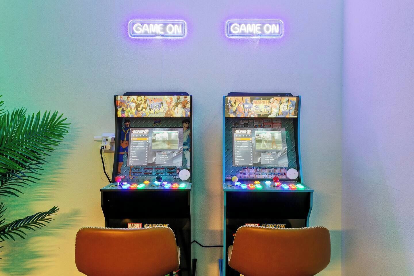 Two arcade gaming machines with neon 'GAME ON' signs, a plant, and a brown chair.