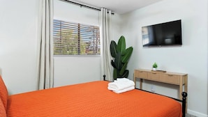 Un dormitorio con cama, televisor, ventana con cortinas, planta y mesita de noche.