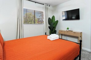 Een slaapkamer met een bed, een televisie, een raam met gordijnen, een plant en een nachtkastje.