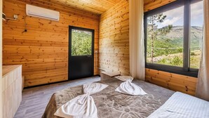 A wooden room with a bed, a window with a view of greenery, and a door.