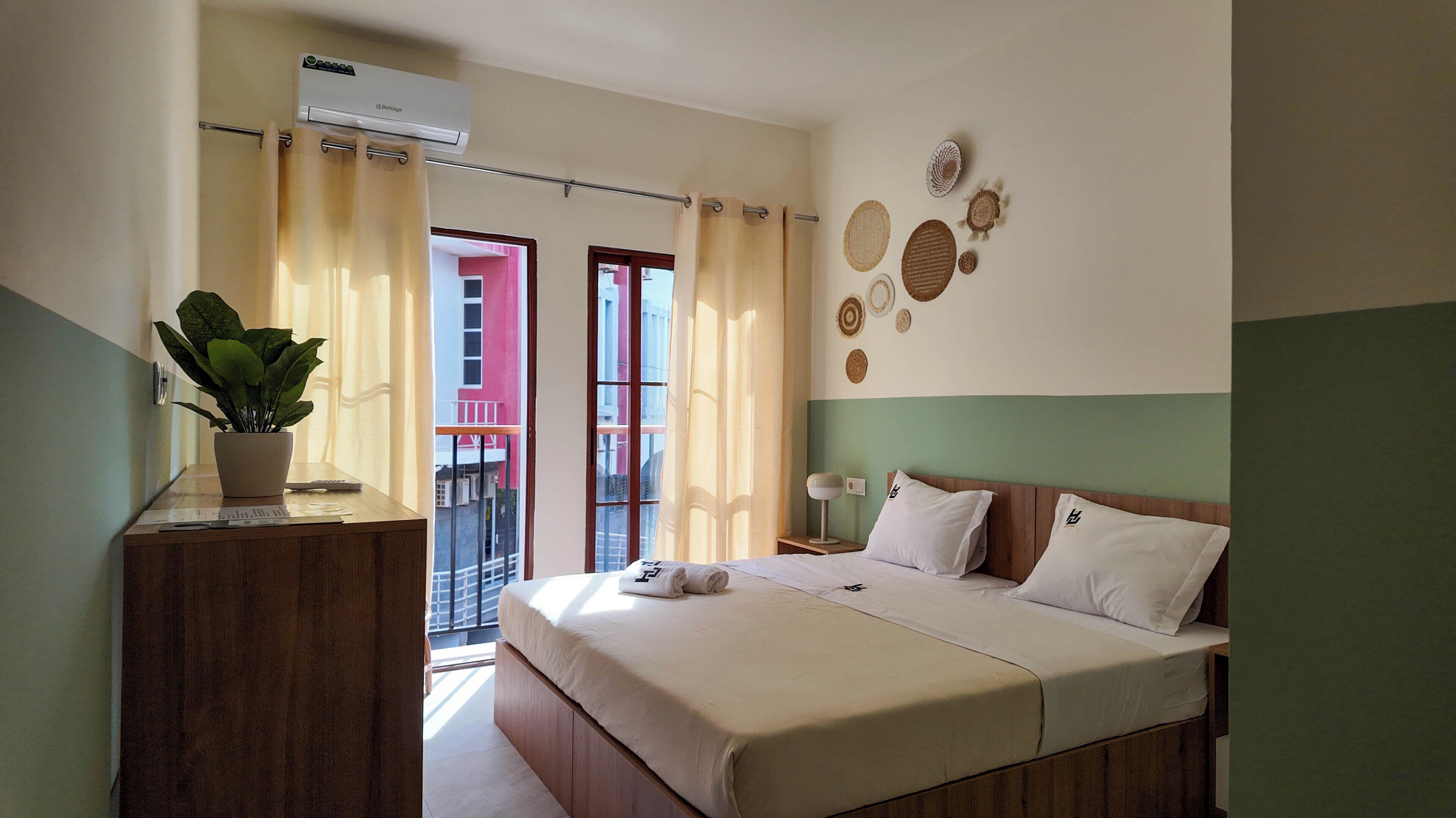Une chambre d’hôtel avec un lit, une table de chevet, une plante en pot, un climatiseur fixé au mur et une vue sur le balcon extérieur.