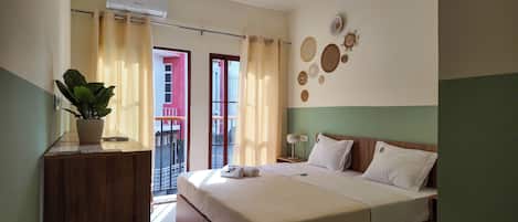 Habitación de hotel con cama, mesita de noche, planta en maceta, aire acondicionado de pared y vistas al balcón exterior.
