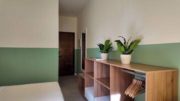 A hotel room with a bed, a wooden console table with a potted plant, and a wooden wardrobe with hanging clothes.