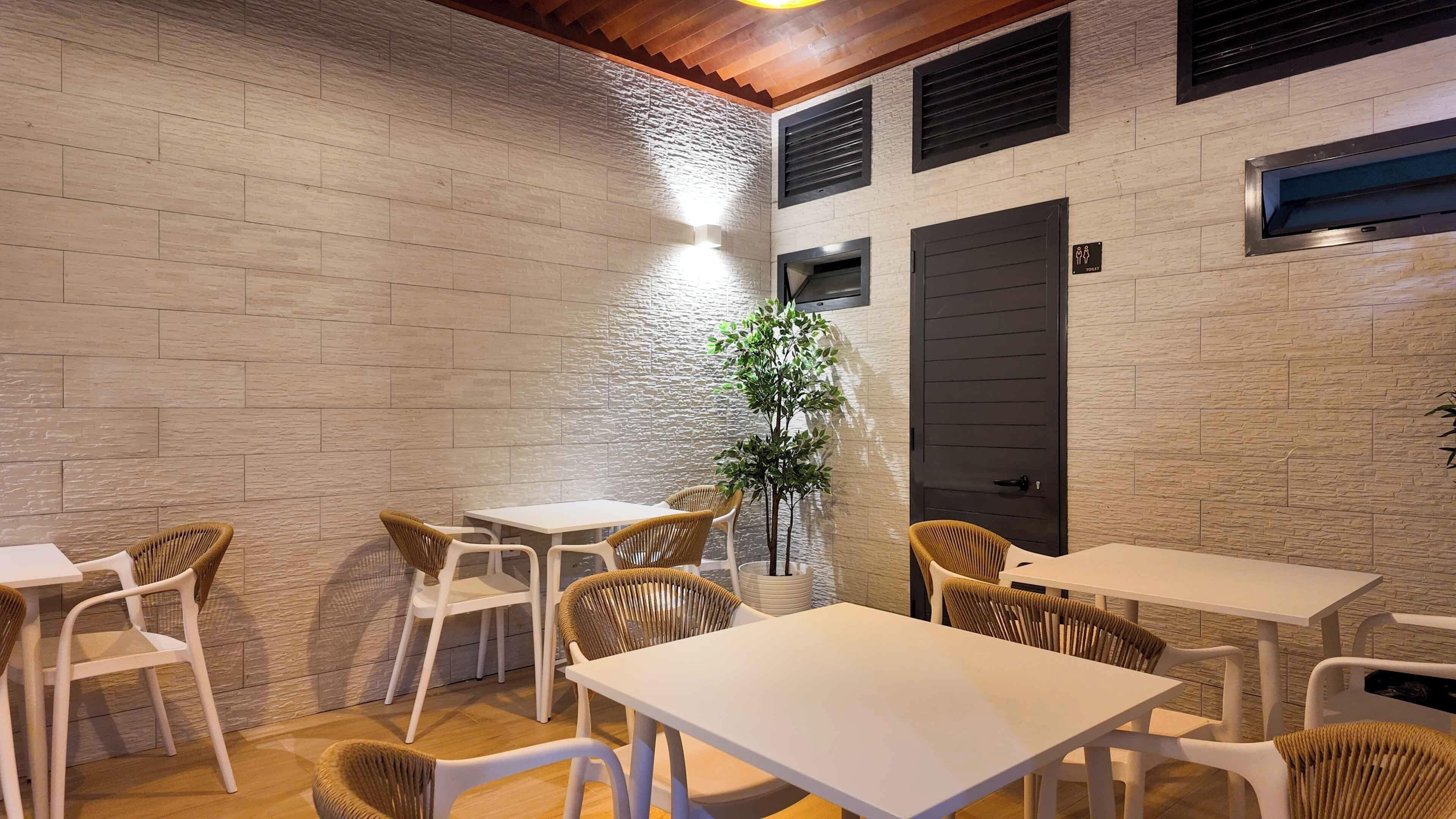 Un espace extérieur moderne, aménagé avec des tables blanches et des chaises en osier, un pot de fleurs et une bouche d’aération fixée au mur.