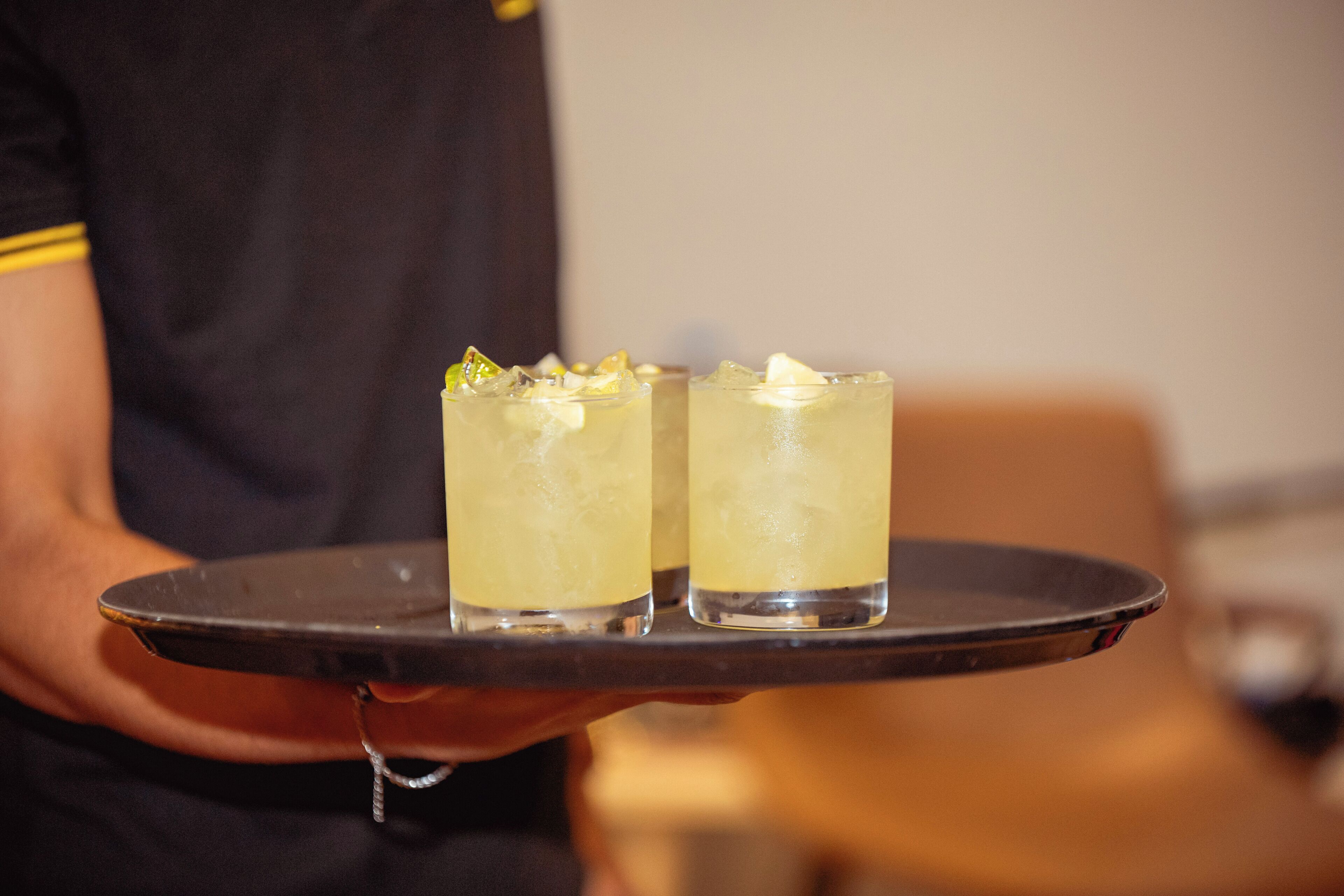 A person holding a tray with two glasses of a yellow beverage, possibly a cocktail, with ice and garnishes.