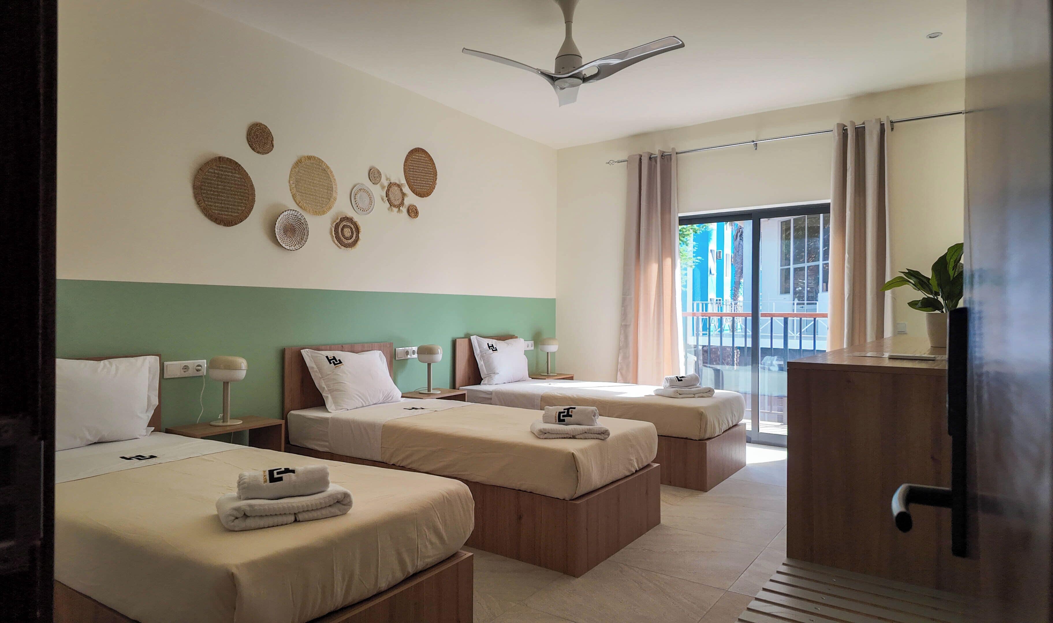 Habitación de hotel con dos camas, un ventilador de techo, un ventanal con cortinas y una planta en maceta.
