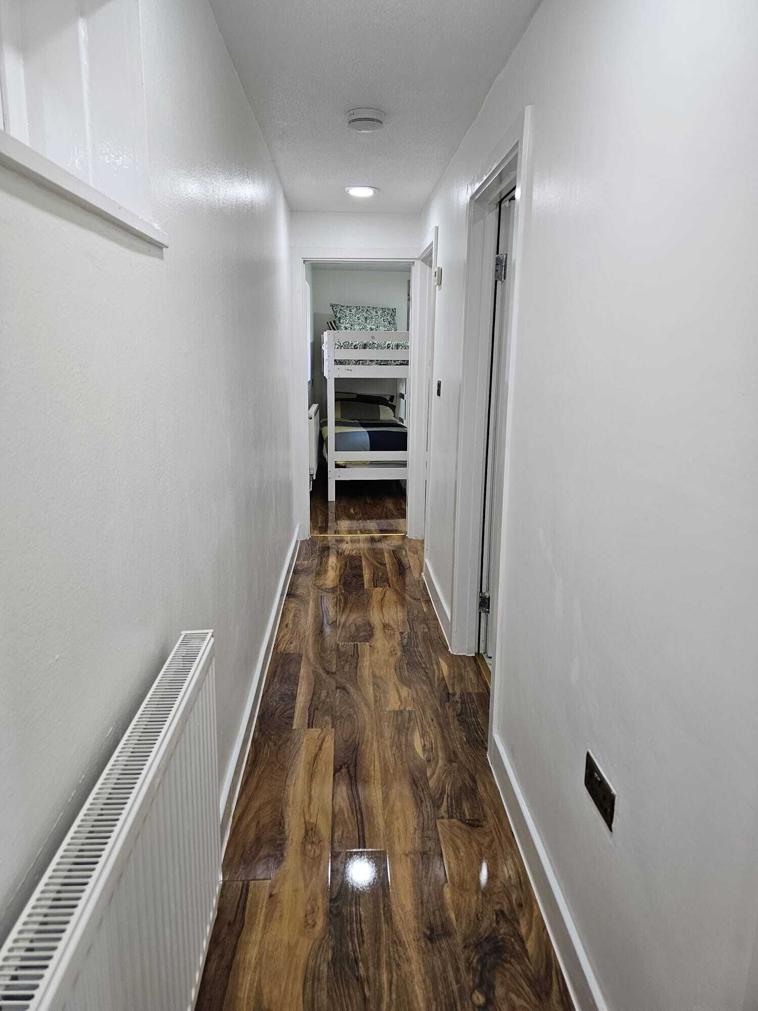 Un couloir étroit avec un plancher en bois, des murs blancs et, au fond, un lit superposé.