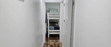 Un couloir étroit avec un plancher en bois, des murs blancs et, au fond, un lit superposé.