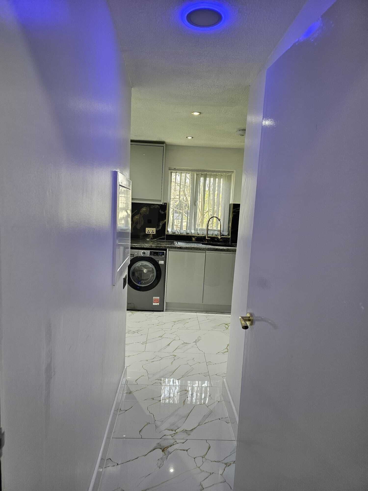 A hallway leading to a kitchen with a washing machine, a window with blinds, and a marble floor.