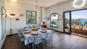 Ein Essbereich mit einem für sechs Personen gedeckten Tisch, einem Deckenventilator und Blick auf das Meer durch eine Glastür.