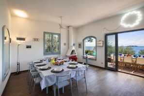 Ein Essbereich mit einem für sechs Personen gedeckten Tisch, einem Deckenventilator und Blick auf das Meer durch eine Glastür.