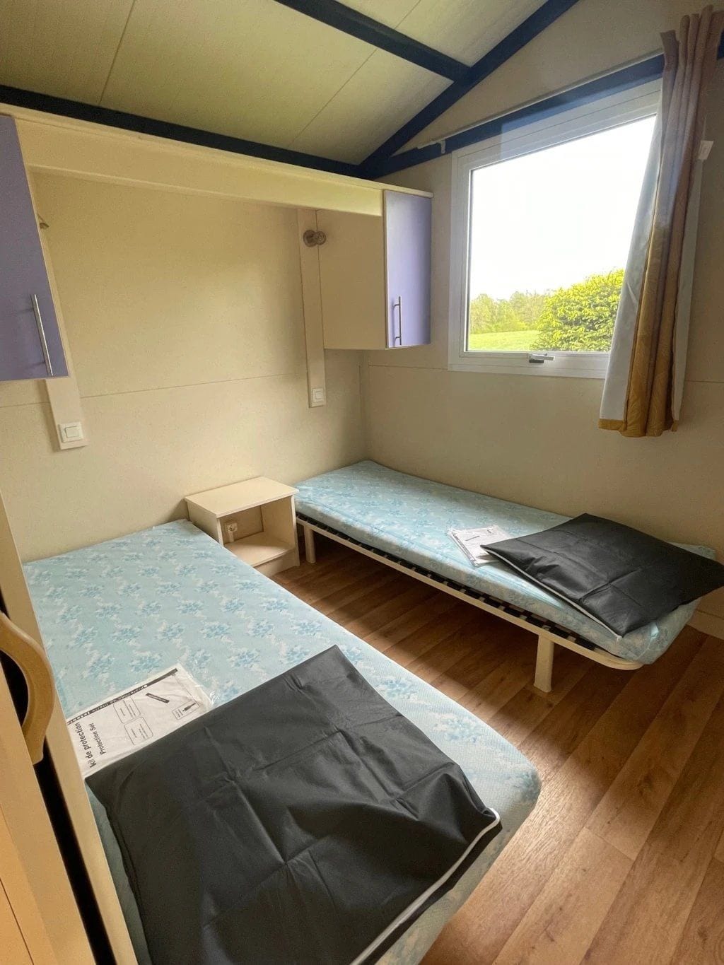 Een knus kamertje met twee bedden, een klein tafeltje, een kast en een raam met gordijnen.