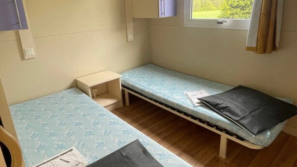 A compact room with two beds, a small table, a cabinet, and a window with curtains. - Chalet 3 étoiles - Piscine  - ccbgahi (Chatel-Chéhéry)