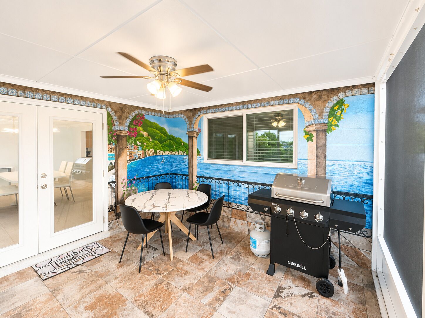 A patio area with a round table, chairs, a grill, and a mural of a coastal scene.