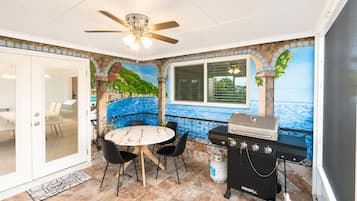 A patio area with a round table, chairs, a grill, and a mural of a coastal scene.